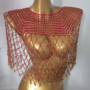 May include: A red crocheted shawl with a diamond net pattern and gold-coloured beads. The shawl is displayed on a gold mannequin. The edges of the shawl are adorned with dangling gold beads.