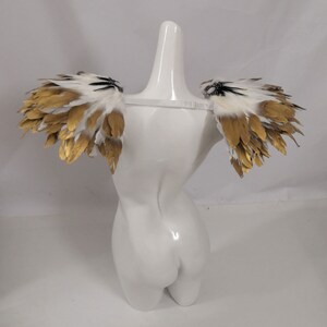 Puede incluir: Un par de alas de hombro de plumas blancas y doradas con detalles negros. Las alas est&aacute;n sujetas a un maniqu&iacute; blanco.