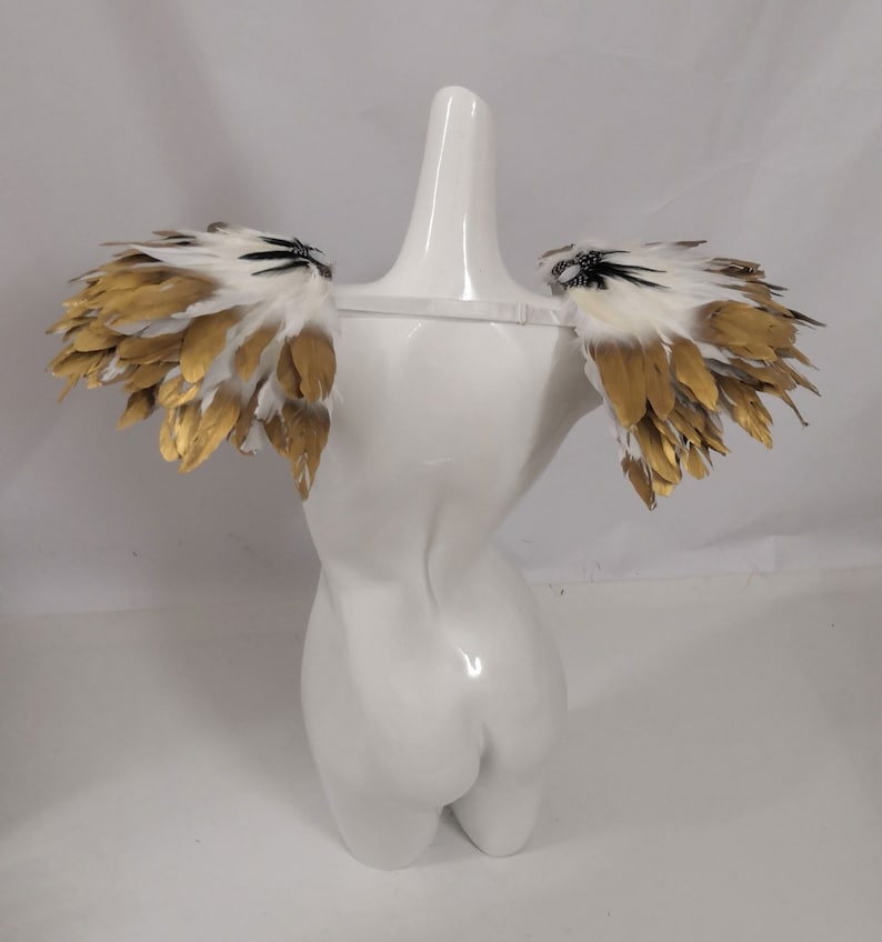 Puede incluir: Un par de alas de hombro de plumas blancas y doradas con detalles negros. Las alas est&aacute;n sujetas a un maniqu&iacute; blanco.