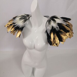Puede incluir: Un par de hombreras de plumas negras y blancas con detalles dorados. Las plumas est&aacute;n dispuestas en forma de abanico en capas.