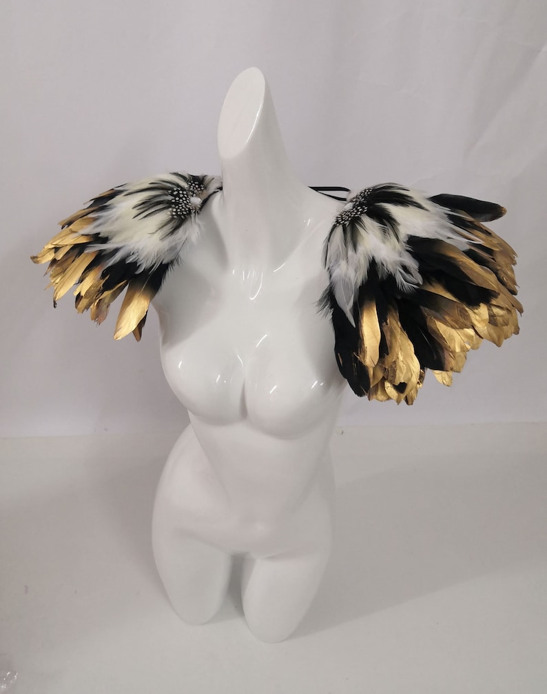 Puede incluir: Un par de hombreras de plumas negras y blancas con detalles dorados. Las plumas est&aacute;n dispuestas en forma de abanico en capas.