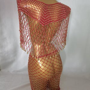 May include: A red beaded net dress and shawl set. The dress is a form-fitting, open-weave design, adorned with small gold beads. The shawl drapes over the shoulders, also featuring gold bead accents. The ensemble is displayed on a mannequin.