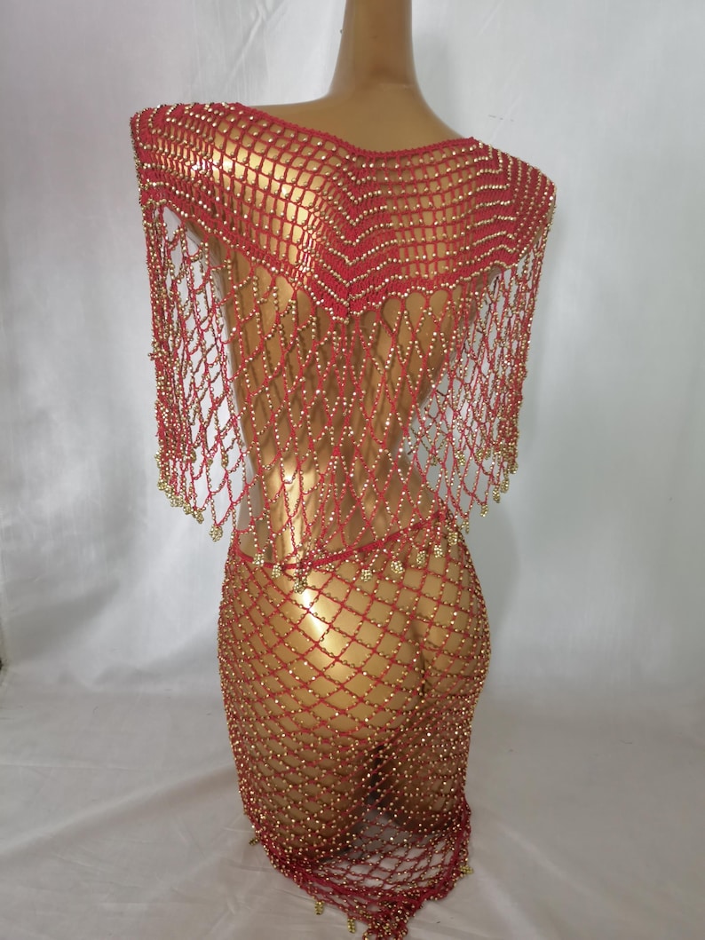 May include: A red beaded net dress and shawl set. The dress is a form-fitting, open-weave design, adorned with small gold beads. The shawl drapes over the shoulders, also featuring gold bead accents. The ensemble is displayed on a mannequin.