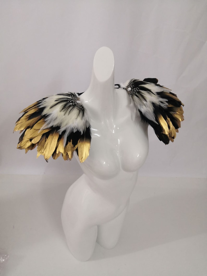 Puede incluir: Un par de hombreras de plumas negras, blancas y doradas con un acento de joya. Las hombreras est&aacute;n en un maniqu&iacute; blanco.