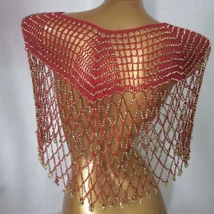 May include: A red, beaded shoulder wrap with a net-like design. The wrap is decorated with small, gold-coloured beads throughout the netting and along the fringe. The garment is displayed on a mannequin.