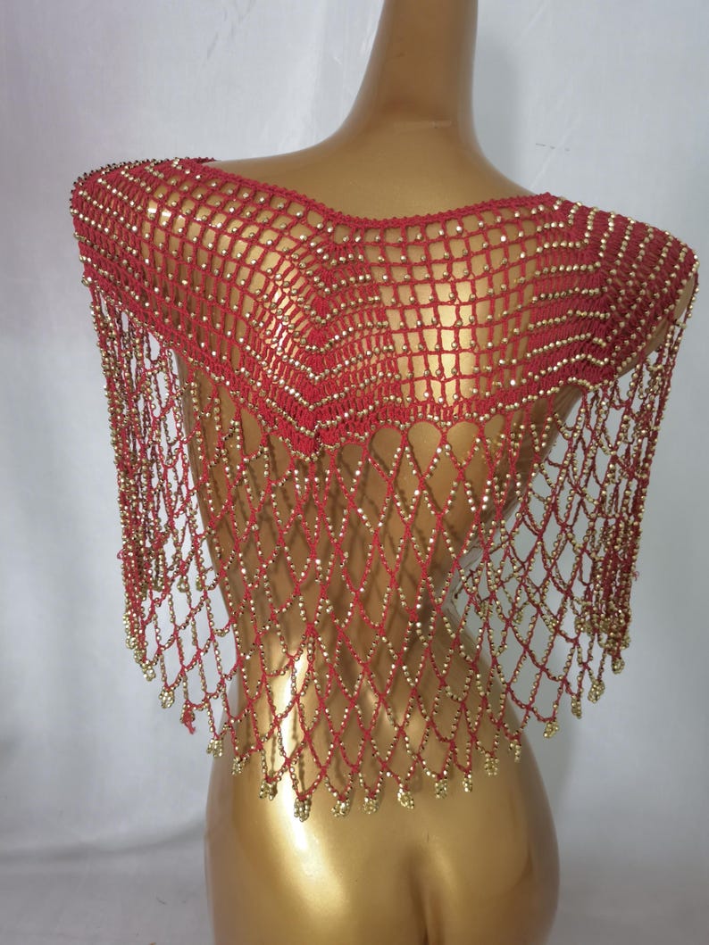 May include: A red, beaded shoulder wrap with a net-like design. The wrap is decorated with small, gold-coloured beads throughout the netting and along the fringe. The garment is displayed on a mannequin.