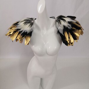 Puede incluir: Un par de hombreras de plumas blancas y negras con detalles dorados. Las hombreras est&aacute;n unidas a una correa negra.