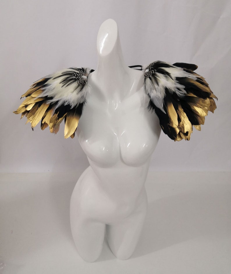Puede incluir: Un par de hombreras de plumas blancas y negras con detalles dorados. Las hombreras est&aacute;n unidas a una correa negra.