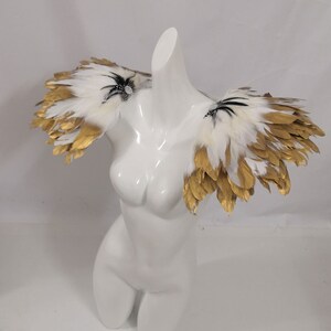Puede incluir: Un par de hombreras de plumas blancas y doradas con detalles negros. Las plumas est&aacute;n dispuestas en un dise&ntilde;o en capas y en cascada.