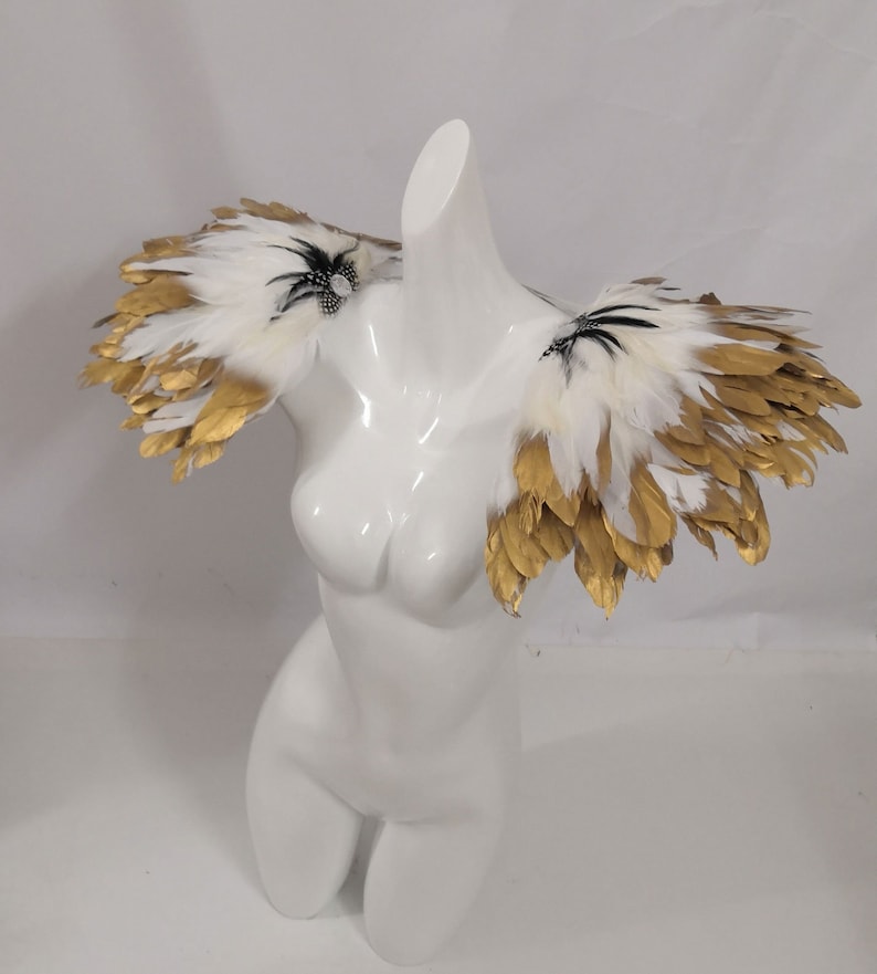 Puede incluir: Un par de hombreras de plumas blancas y doradas con detalles negros. Las plumas est&aacute;n dispuestas en un dise&ntilde;o en capas y en cascada.