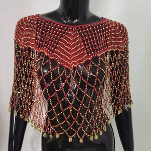 May include: A red beaded shawl with a net-like design. The shawl features gold-coloured beads and a fringe along the bottom edge. The garment is displayed on a black mannequin.