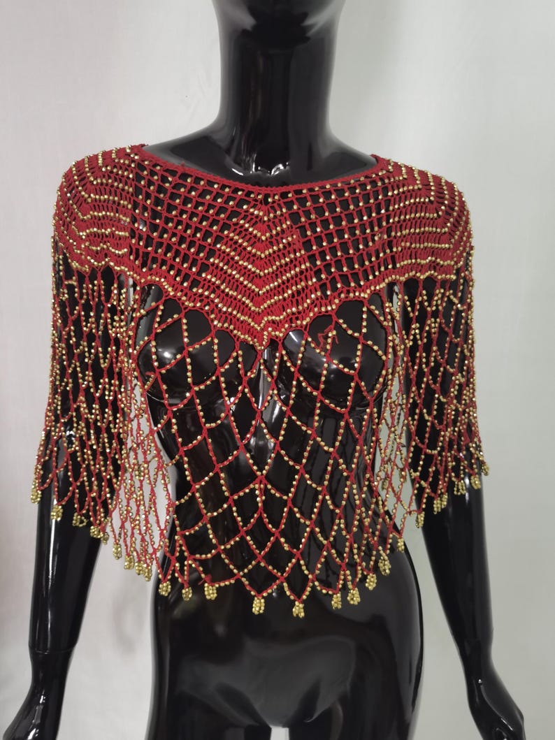 May include: A red beaded shawl with a net-like design. The shawl features gold-coloured beads and a fringe along the bottom edge. The garment is displayed on a black mannequin.