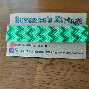 Può includere: Braccialetto di amicizia a chevron verde e bianco su una carta marrone con il testo "Suzanne's Strings" e informazioni di contatto.