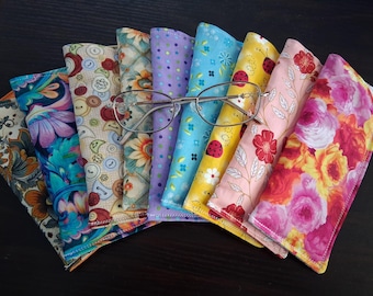 May include: A collection of eight fabric eyeglass cases arranged in a fan shape, featuring diverse patterns including floral prints, polka dots, buttons, and ladybugs. A pair of thin-framed metal eyeglasses rests horizontally across the center.