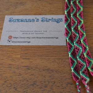 Pu&ograve; includere: Quattro braccialetti dell'amicizia colorati con un motivo a cocomero in rosso, verde, nero e giallo. I braccialetti sono legati insieme e sono su una superficie di legno. Il testo sulla carta dice "Suzanne's Strings" e "Friendship makes the world go 'round!" con un sito web e un handle di social media.