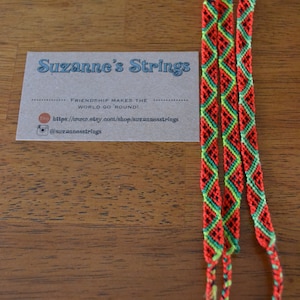 Pu&ograve; includere: Tre braccialetti dell'amicizia rossi, verdi e neri con un motivo a rombi. I braccialetti sono su una superficie di legno con un biglietto da visita che recita "Suzanne's Strings" e "Friendship makes the world go 'round!" con un sito web e un account Instagram.