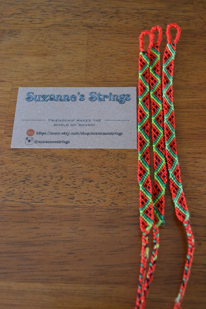 Pu&ograve; includere: Tre braccialetti dell'amicizia rossi, verdi e neri con un motivo a rombi. I braccialetti sono su una superficie di legno con un biglietto da visita che recita "Suzanne's Strings" e "Friendship makes the world go 'round!" con un sito web e un account Instagram.