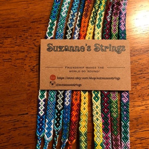 Pode incluir: Uma cole&ccedil;&atilde;o de 14 pulseiras de amizade coloridas com v&aacute;rios padr&otilde;es geom&eacute;tricos. As pulseiras s&atilde;o exibidas em um cart&atilde;o marrom com o texto "Suzanne's Strings" e o slogan "A amizade faz o mundo girar!"