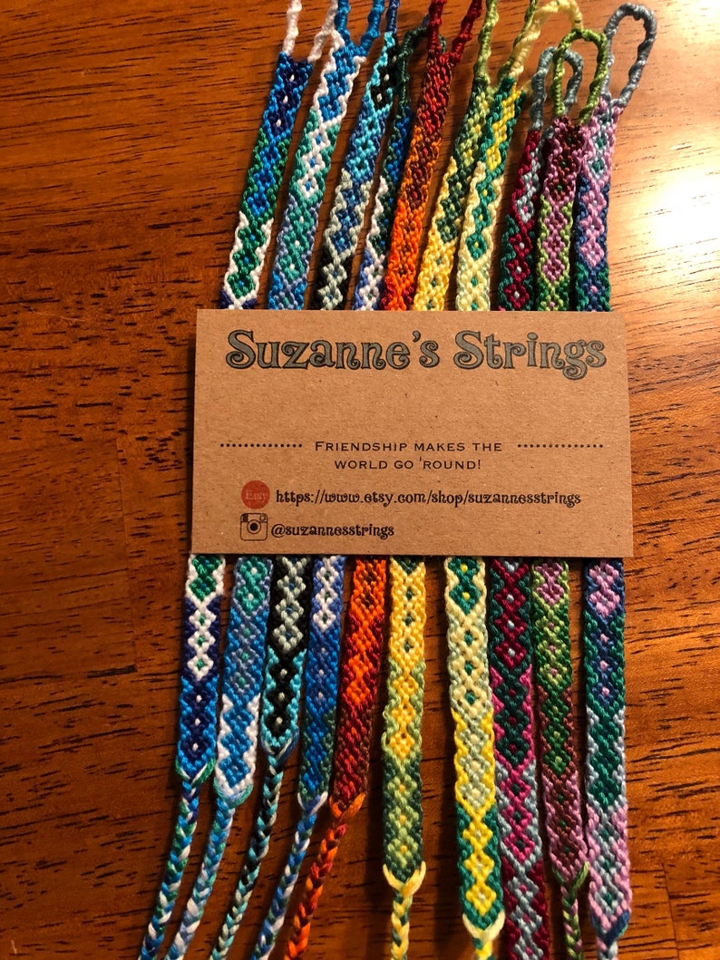 Pode incluir: Uma cole&ccedil;&atilde;o de 14 pulseiras de amizade coloridas com v&aacute;rios padr&otilde;es geom&eacute;tricos. As pulseiras s&atilde;o exibidas em um cart&atilde;o marrom com o texto "Suzanne's Strings" e o slogan "A amizade faz o mundo girar!"