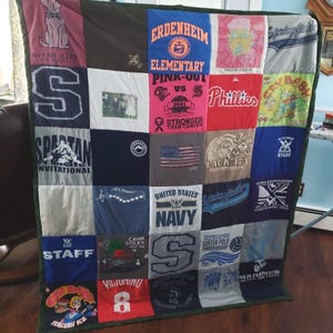 May include: A patchwork quilt made from various t-shirts, featuring logos, text, and graphics. The quilt includes a variety of colors and designs, with a green border. The quilt is approximately 60 inches by 70 inches.