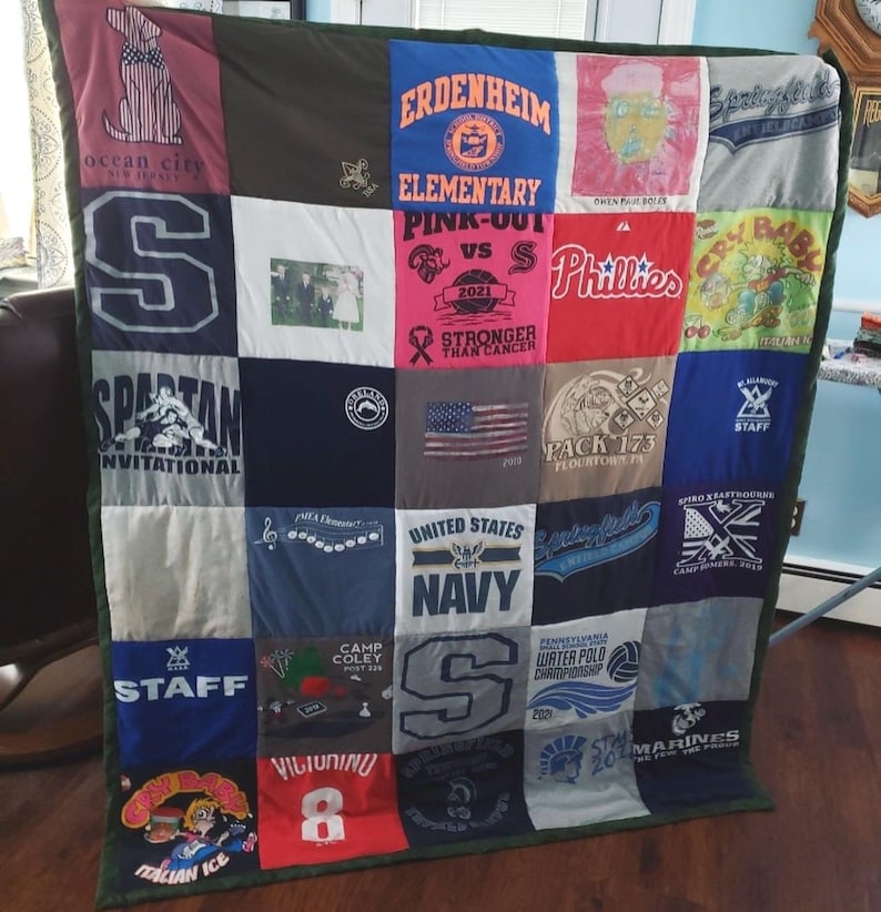 May include: A patchwork quilt made from various t-shirts, featuring logos, text, and graphics. The quilt includes a variety of colours and designs, with a green border. The quilt is approximately 152 cm by 178 cm.