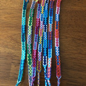 Pode incluir: Sete pulseiras de amizade coloridas com padr&otilde;es e cores diferentes, incluindo azul, verde, amarelo, rosa, laranja e branco.
