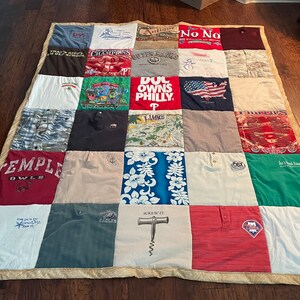 May include: A patchwork quilt constructed from repurposed t-shirts, showcasing a diverse array of colours and graphics. The quilt incorporates shirts with text such as "TEMPLE OWLS", "EAGLES", and "SCREW IT". It is edged with a patterned fabric.