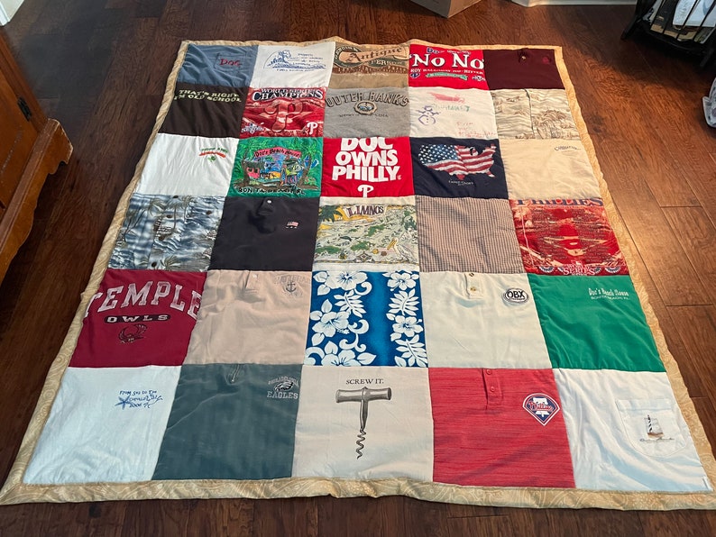 May include: A patchwork quilt constructed from repurposed t-shirts, showcasing a diverse array of colours and graphics. The quilt incorporates shirts with text such as "TEMPLE OWLS", "EAGLES", and "SCREW IT". It is edged with a patterned fabric.