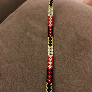 Pode incluir: Um bracelete de amizade preto, branco, vermelho e amarelo com um padr&atilde;o de diamante.