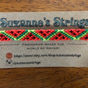 Pu&ograve; includere: Un braccialetto dell'amicizia rosso, verde e nero con un motivo a cocomero. Il braccialetto &egrave; su una carta marrone con il testo "Suzanne's Strings" e "Friendship makes the world go 'round!" La carta include anche un link al negozio Etsy e un nome utente Instagram.