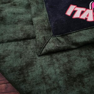 May include: Dark green fabric with a textured pattern, featuring a black corner with the word "ITA" in red and white. The fabric has a quilted appearance.