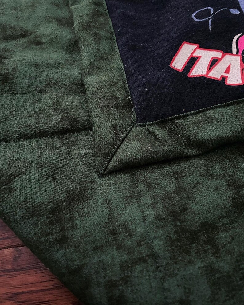 May include: Dark green fabric with a textured pattern, featuring a black corner with the word "ITA" in red and white. The fabric has a quilted appearance.
