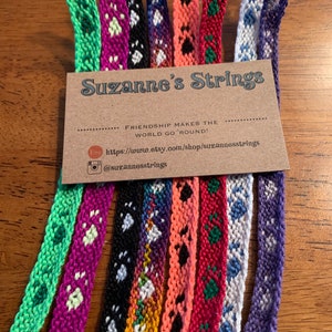 May include: A collection of colorful friendship bracelets with various patterns, including paw prints, hearts, and geometric designs. The bracelets are displayed on a brown card with the text "Suzanne's Strings" and the tagline "Friendship makes the world go 'round!"
