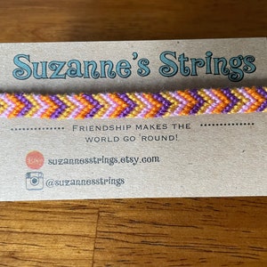 May include: A colorful friendship bracelet with orange, purple, pink, and yellow threads woven in a chevron pattern. The bracelet is displayed on a card with the text "Suzanne's Strings" and "Friendship makes the world go 'round!"