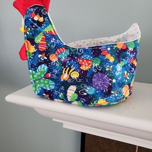May include: A blue fabric chicken-shaped basket with a red comb and a white lining. The basket is decorated with a colorful pattern of bugs, butterflies, and stars.