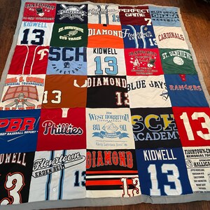 May include: A patchwork quilt made from sports team jerseys and t-shirts. The quilt features a variety of colours, team names, and numbers, including "13" and "Phillies". The quilt is bordered with a grey fabric. The quilt is approximately 180 cm x 180 cm.