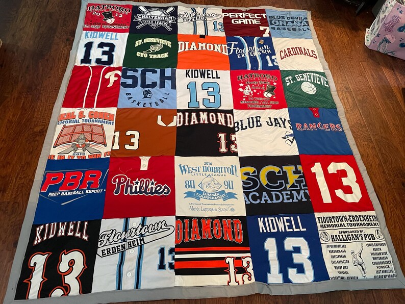May include: A patchwork quilt made from sports team jerseys and t-shirts. The quilt features a variety of colours, team names, and numbers, including "13" and "Phillies". The quilt is bordered with a grey fabric. The quilt is approximately 180 cm x 180 cm.