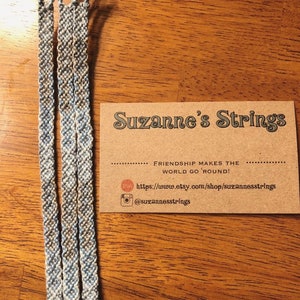 Pode incluir: Tr&ecirc;s pulseiras de amizade azul e branca com um cart&atilde;o castanho que diz "Suzanne's Strings" e "Friendship makes the world go 'round!" com um site e um nome de utilizador do Instagram.