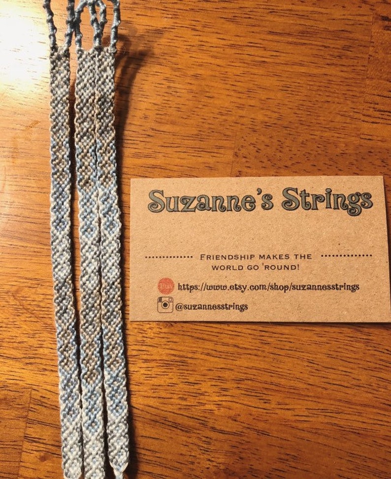Pode incluir: Tr&ecirc;s pulseiras de amizade azul e branca com um cart&atilde;o castanho que diz "Suzanne's Strings" e "Friendship makes the world go 'round!" com um site e um nome de utilizador do Instagram.
