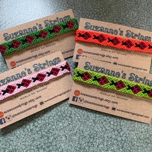 May include: Four woven friendship bracelets displayed on individual kraft paper cards labeled Suzanne's Strings. Each bracelet features a repeating diamond pattern in contrasting colors, including green, red, orange, pink, and black thread designs.