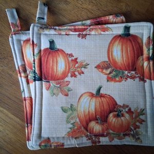 May include: Two square quilted pot holders with a fall theme. The beige fabric is printed with orange pumpkins, green stems, and autumn leaves. Each pot holder has a loop for hanging and a cream-colored border.