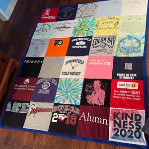 May include: A patchwork quilt made from repurposed t-shirts, each square showcasing different designs and text. The quilt, edged with blue fabric, lies on a wooden floor. The t-shirts feature diverse colours and graphics, creating a vibrant textile.