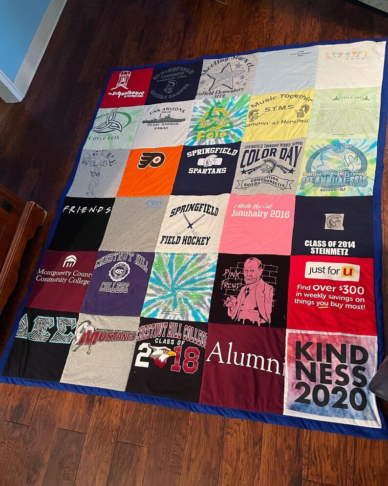 May include: A patchwork quilt made from repurposed t-shirts, each square showcasing different designs and text. The quilt, edged with blue fabric, lies on a wooden floor. The t-shirts feature diverse colours and graphics, creating a vibrant textile.