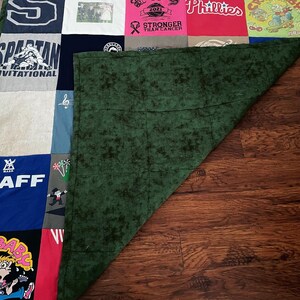 May include: A patchwork quilt made from repurposed t-shirts, displaying a variety of logos, text, and graphics. The quilt features a mix of colours, including red, blue, and green, with a dark green backing. The quilt is displayed on a wooden surface.