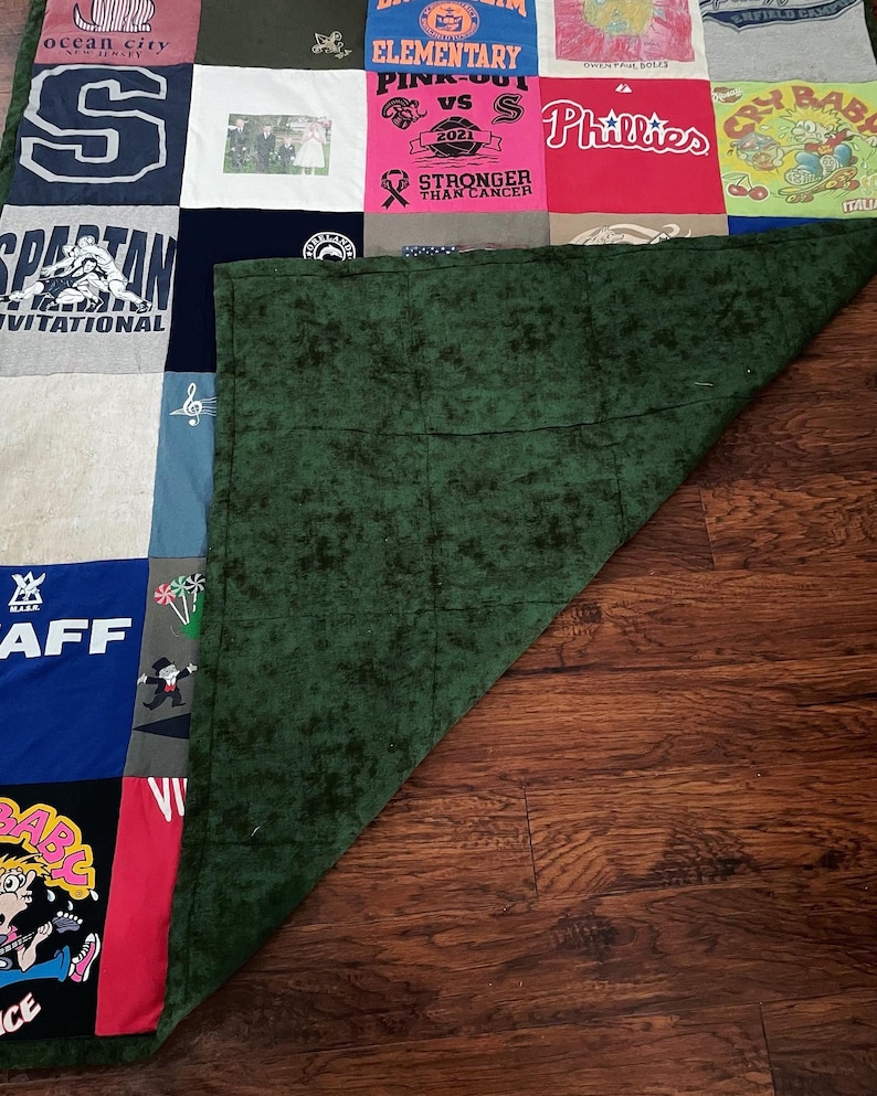 May include: A patchwork quilt made from repurposed t-shirts, displaying a variety of logos, text, and graphics. The quilt features a mix of colours, including red, blue, and green, with a dark green backing. The quilt is displayed on a wooden surface.