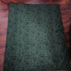 May include: A dark green quilt with a mottled pattern, laid out on a wooden floor. The quilt is divided into rectangular sections by stitched lines. The fabric has a textured appearance, with darker green and black accents throughout.