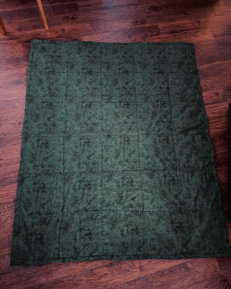 May include: A dark green quilt with a mottled pattern, laid out on a wooden floor. The quilt is divided into rectangular sections by stitched lines. The fabric has a textured appearance, with darker green and black accents throughout.