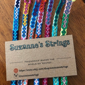 Pode incluir: Um close-up de oito pulseiras de amizade coloridas com um cart&atilde;o marrom que diz "Suzanne's Strings" e "Friendship makes the world go 'round!" com um site e uma al&ccedil;a de m&iacute;dia social.