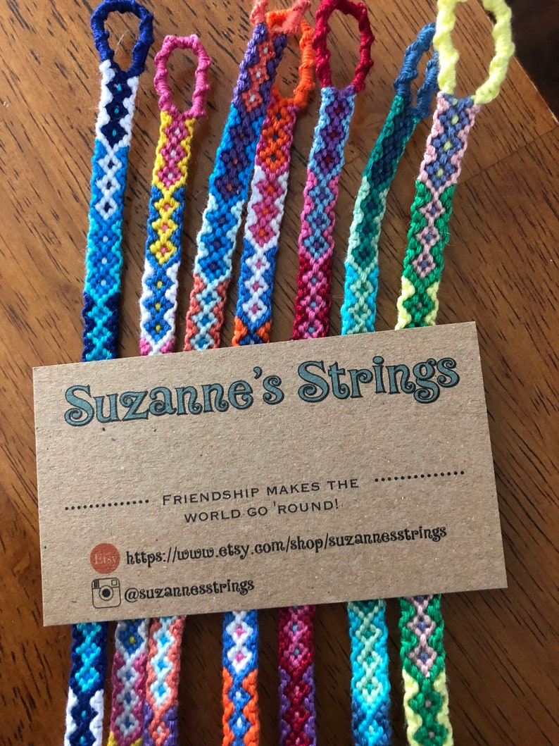Pode incluir: Um close-up de oito pulseiras de amizade coloridas com um cart&atilde;o marrom que diz "Suzanne's Strings" e "Friendship makes the world go 'round!" com um site e uma al&ccedil;a de m&iacute;dia social.