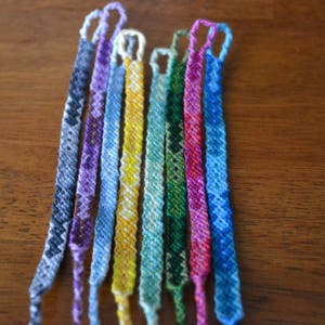 Pode incluir: Uma cole&ccedil;&atilde;o de dez pulseiras de amizade coloridas, cada uma com um padr&atilde;o &uacute;nico. As pulseiras s&atilde;o feitas com diferentes tons de azul, roxo, verde, amarelo e vermelho.